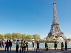Reimagined Landmarks and Experiences Across the Paris Region Tourists take photos of the Eiffel Tower from the banks of the Seine River in Paris, France, under a clear blue sky with riverboats and trees lining the waterfront.