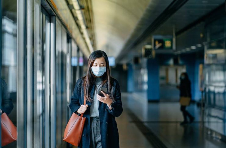 Asian woman with protective face mask using smartphone while walking on the subway platform