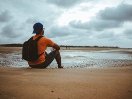 Man in Orange Shirt with Backpack Sitting on Beach Sand