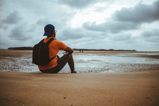 Man in Orange Shirt with Backpack Sitting on Beach Sand