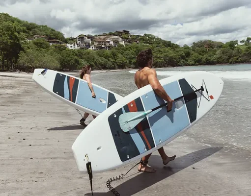 Blue Zone Inspired Living at Las Catalinas in Costa Rica Two individuals are walking along a sandy beach, each carrying a surfboard with blue and red designs.