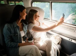 Two women sitting inside a train compartment. One woman, wearing a denim jacket, is looking at the other woman who is holding a smartphone and taking a selfie. The woman with the smartphone is dressed in a white outfit. They are seated next to a large window through which trees can be seen outside. On the seat beside them, there's a coffee cup and a white cloth item.