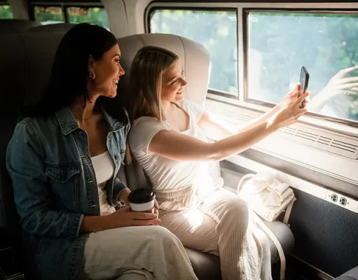 Amtrak Customers Now Benefit From More Flexible and Affordable Fares Two women sitting inside a train compartment. One woman, wearing a denim jacket, is looking at the other woman who is holding a smartphone and taking a selfie. The woman with the smartphone is dressed in a white outfit. They are seated next to a large window through which trees can be seen outside. On the seat beside them, there's a coffee cup and a white cloth item.