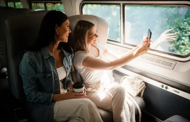 Two women sitting inside a train compartment. One woman, wearing a denim jacket, is looking at the other woman who is holding a smartphone and taking a selfie. The woman with the smartphone is dressed in a white outfit. They are seated next to a large window through which trees can be seen outside. On the seat beside them, there's a coffee cup and a white cloth item.