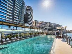 Barceló Hotel Group Opens a 5-star Hotel in Malta An urban waterfront setting with a clear blue swimming pool adjacent to modern high-rise buildings. The pool is lined with white loungers and shaded by large umbrellas. The background features a mix of contemporary architecture with glimpses of the water beyond. The sun shines brightly in the clear sky, casting reflections on the water's surface.