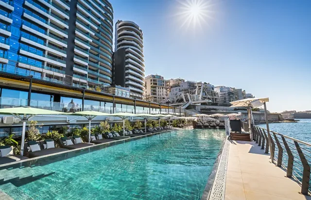 An urban waterfront setting with a clear blue swimming pool adjacent to modern high-rise buildings. The pool is lined with white loungers and shaded by large umbrellas. The background features a mix of contemporary architecture with glimpses of the water beyond. The sun shines brightly in the clear sky, casting reflections on the water's surface.