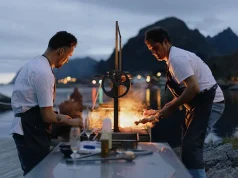 Acclaimed chefs join Holmen Lofoten for Kitchen On The Edge 2024 for a remarkable programme of adventures on Norway’s remote Lofoten archipelago Two chefs cooking outdoors by a waterfront, with a fiery grill illuminating them against a backdrop of mountains and a twilight sky.