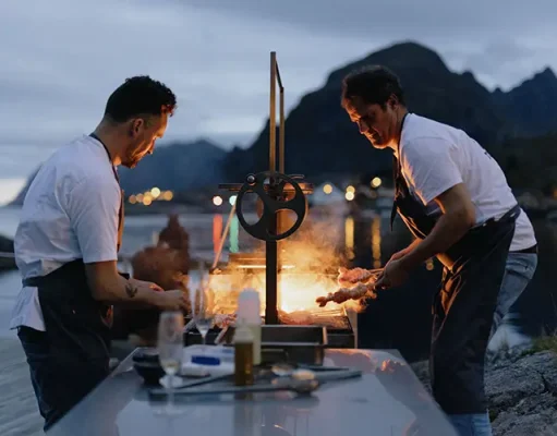 Acclaimed chefs join Holmen Lofoten for Kitchen On The Edge 2024 for a remarkable programme of adventures on Norway’s remote Lofoten archipelago Two chefs cooking outdoors by a waterfront, with a fiery grill illuminating them against a backdrop of mountains and a twilight sky.