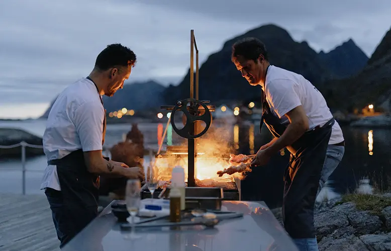 Two chefs cooking outdoors by a waterfront, with a fiery grill illuminating them against a backdrop of mountains and a twilight sky.