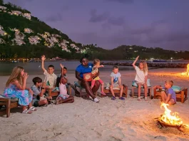 A group of diverse children sitting on beach chairs around a fire, listening intently to a woman reading a book, with a backdrop of a twilight beach scene and illuminated cliffs.