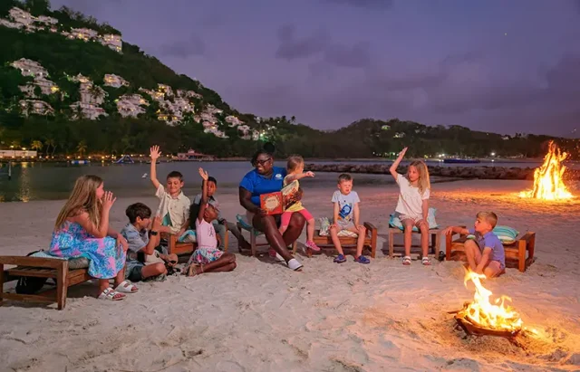 A group of diverse children sitting on beach chairs around a fire, listening intently to a woman reading a book, with a backdrop of a twilight beach scene and illuminated cliffs.