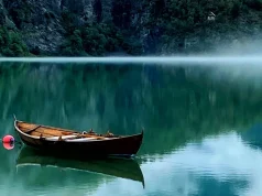 Up Norway Announces New Wittgenstein-Inspired Workcation for Creatives Wooden boat anchored on a calm, reflective green lake with mist hovering above the water, steep rocky cliffs in the background and a distant cabin nestled among trees
