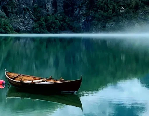 Up Norway Announces New Wittgenstein-Inspired Workcation for Creatives Wooden boat anchored on a calm, reflective green lake with mist hovering above the water, steep rocky cliffs in the background and a distant cabin nestled among trees