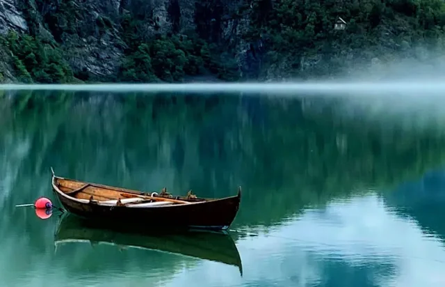 Wooden boat anchored on a calm, reflective green lake with mist hovering above the water, steep rocky cliffs in the background and a distant cabin nestled among trees