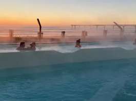 People relaxing in an outdoor infinity hot tub with steam rising from the water, overlooking a serene sunset with a clear sky. In the background, there's a silhouette of a pier extending into the calm sea
