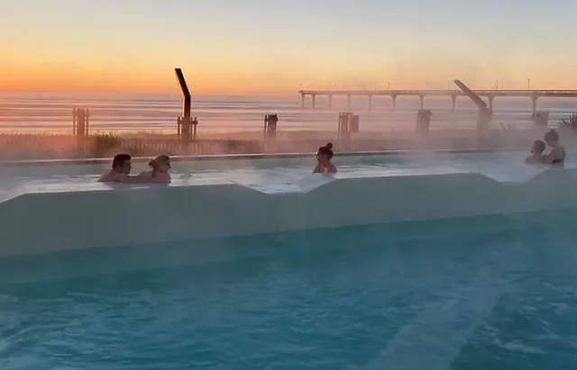 People relaxing in an outdoor infinity hot tub with steam rising from the water, overlooking a serene sunset with a clear sky. In the background, there's a silhouette of a pier extending into the calm sea