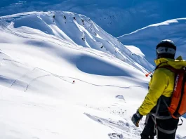 A snowboarder in a bright yellow jacket and black helmet with an orange backpack stands facing a vast snow-covered mountain landscape. In the middle ground, another snowboarder can be seen carving a trail down the pristine slopes. The sky is a clear, deep blue, indicating a perfect day for snowboarding with excellent visibility.