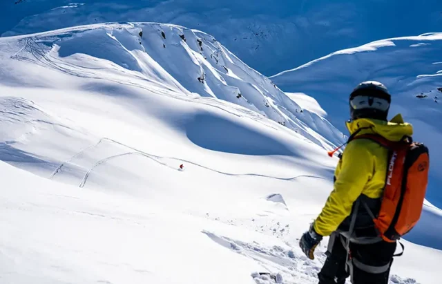 A snowboarder in a bright yellow jacket and black helmet with an orange backpack stands facing a vast snow-covered mountain landscape. In the middle ground, another snowboarder can be seen carving a trail down the pristine slopes. The sky is a clear, deep blue, indicating a perfect day for snowboarding with excellent visibility.