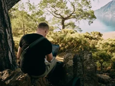 Traveling as a Digital Nomad: Work and Explore the World man sitting on a rock working on his laptop. Mountain and lake in the background.