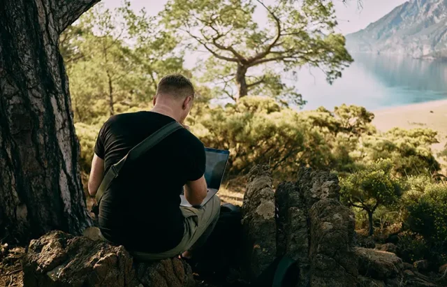 man sitting on a rock working on his laptop. Mountain and lake in the background.