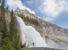 Essential Tips for Acquiring an ETA and Exploring Canada A person stands in awe at the base of a majestic waterfall cascading down a sheer cliff face surrounded by lush greenery and rugged mountain peaks under a partly cloudy sky.