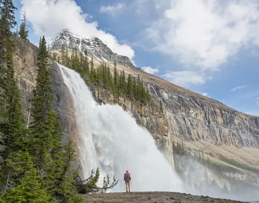 Essential Tips for Acquiring an ETA and Exploring Canada A person stands in awe at the base of a majestic waterfall cascading down a sheer cliff face surrounded by lush greenery and rugged mountain peaks under a partly cloudy sky.