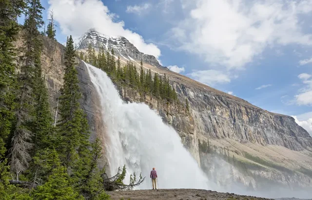 A person stands in awe at the base of a majestic waterfall cascading down a sheer cliff face surrounded by lush greenery and rugged mountain peaks under a partly cloudy sky.