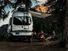 Winter Camping on the Rise: Here are 5 Trips to Try three people relaxes by a campfire in a forest at dusk, with one person playing the guitar. They are seated next to a Winnebago Revel 4x4 RV, which has its awning extended. The surrounding trees and the mountain in the background are bathed in the soft glow of the setting sun.