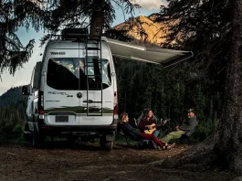 three people relaxes by a campfire in a forest at dusk, with one person playing the guitar. They are seated next to a Winnebago Revel 4x4 RV, which has its awning extended. The surrounding trees and the mountain in the background are bathed in the soft glow of the setting sun.