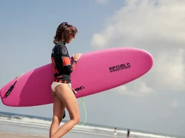 A woman walking on a beach carrying a bright pink surfboard under her arm. The surfboard has the text "RIP CURL SCHOOL OF SURF" printed on it. She's wearing a black short-sleeved wetsuit with red accents and shorts. Her hair is short, and she's looking down as she walks. The ocean can be seen in the background with a few surfers and the sky is partly cloudy.