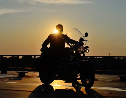 Solo Motorcycle Travel Safety: Tips for Riding Alone silhouette of a man on a motorcycle on the road, behind him the setting sun, warm, yellow tones