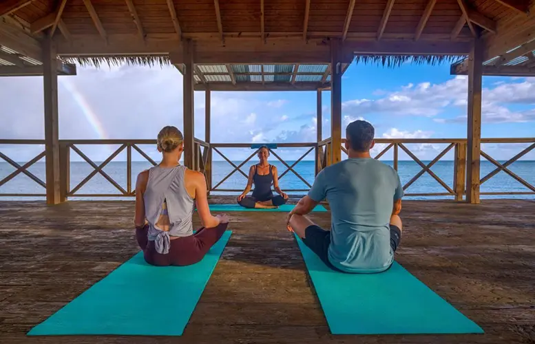 Two participants are seated on yoga mats, engaging in a meditative posture, while an instructor is positioned facing them, guiding the practice. The open structure of the pavilion allows for a panoramic view of the surrounding seascape, and the presence of a rainbow adds a serene and picturesque quality to the setting, suggesting a peaceful retreat or wellness-focused resort.
