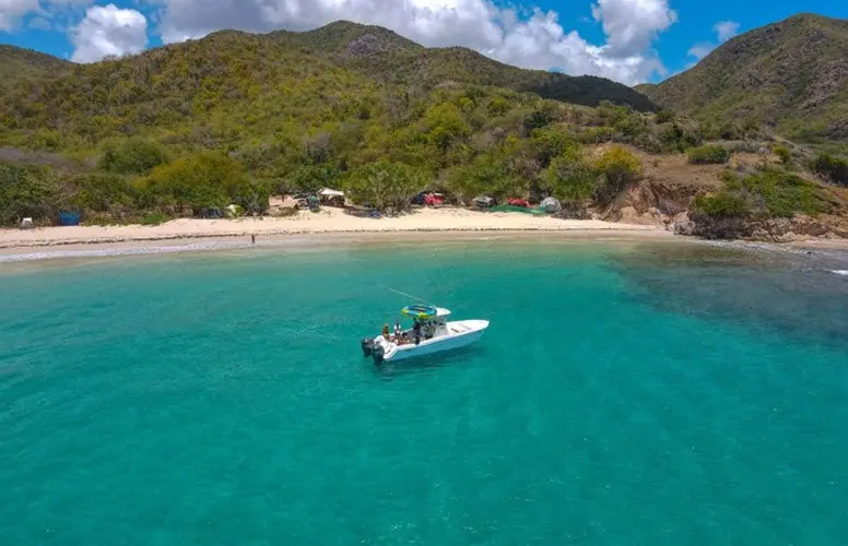 aerial view of a small motorboat on clear turquoise waters near a sandy beach. The beach is fringed by lush greenery and backed by rolling hills. The boat appears to have a few passengers on board, possibly on a tour or a recreational outing. The setting suggests a tropical or subtropical location, ideal for water activities and enjoying the natural beauty of the coastline.