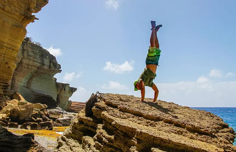 person performing a handstand on rocky terrain against a dramatic backdrop of layered sedimentary cliffs and the ocean. 