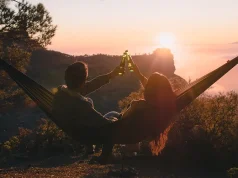 Wanting to travel more in 2024? beautiful couple seated in a hammock toasting with a cloudscape sunset