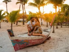 Top Wedding and Honeymoon Spots in St. Martin Revealed A couple sitting closely together on a vibrantly painted boat on a sandy beach with palm trees in the background during sunset