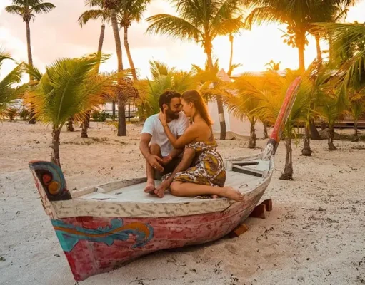 Top Wedding and Honeymoon Spots in St. Martin Revealed A couple sitting closely together on a vibrantly painted boat on a sandy beach with palm trees in the background during sunset