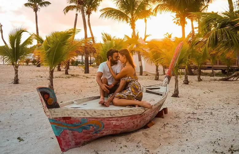 A couple sitting closely together on a vibrantly painted boat on a sandy beach with palm trees in the background during sunset