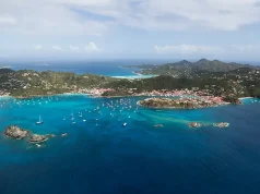Villa Luxury Rental in St. Barth Aerial view of a stunning tropical bay, showcasing vibrant turquoise waters with scattered boats anchored near a shoreline. The coastline is dotted with densely clustered red-roofed buildings, surrounded by lush greenery and rolling hills in the backdrop, under a sky lightly brushed with wispy clouds.