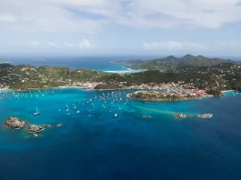 Aerial view of a stunning tropical bay, showcasing vibrant turquoise waters with scattered boats anchored near a shoreline. The coastline is dotted with densely clustered red-roofed buildings, surrounded by lush greenery and rolling hills in the backdrop, under a sky lightly brushed with wispy clouds.