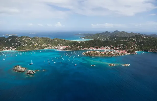 Aerial view of a stunning tropical bay, showcasing vibrant turquoise waters with scattered boats anchored near a shoreline. The coastline is dotted with densely clustered red-roofed buildings, surrounded by lush greenery and rolling hills in the backdrop, under a sky lightly brushed with wispy clouds.
