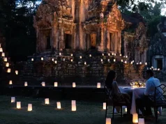 Raffles Grand Hotel d’Angkor Unveils New ‘Curated Journeys’ and ‘Resort Program’ A romantic dinner for two set up at dusk in front of an ancient temple. The area is softly illuminated by numerous small, glowing lanterns placed on the ground, creating a path of light leading to the temple. The couple sits at a table with a white cloth, fine dinnerware, and lit candles, engrossed in conversation.