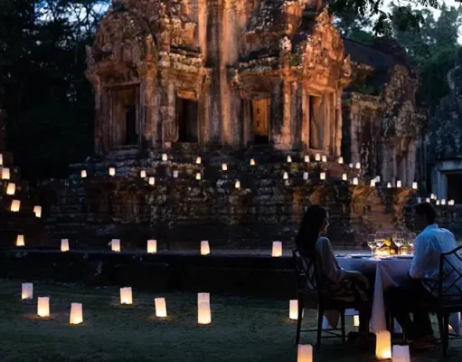 Raffles Grand Hotel d’Angkor Unveils New ‘Curated Journeys’ and ‘Resort Program’ A romantic dinner for two set up at dusk in front of an ancient temple. The area is softly illuminated by numerous small, glowing lanterns placed on the ground, creating a path of light leading to the temple. The couple sits at a table with a white cloth, fine dinnerware, and lit candles, engrossed in conversation.