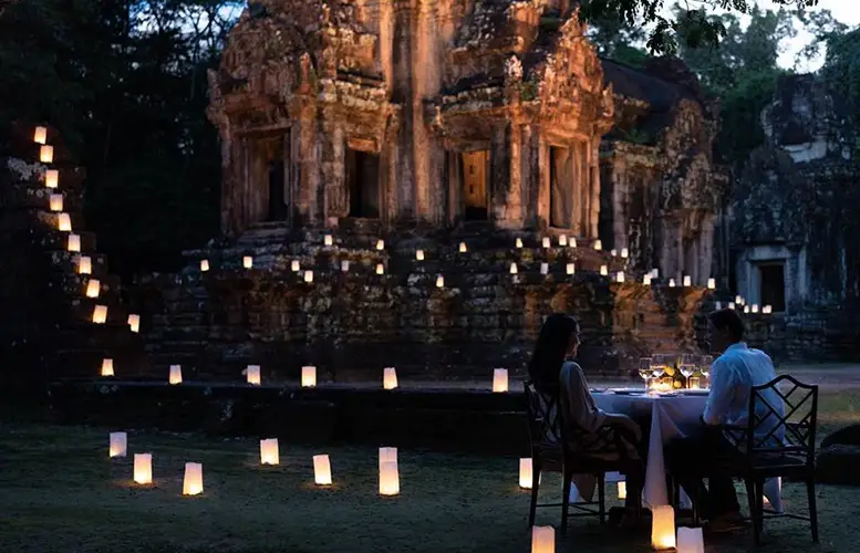 A romantic dinner for two set up at dusk in front of an ancient temple. The area is softly illuminated by numerous small, glowing lanterns placed on the ground, creating a path of light leading to the temple. The couple sits at a table with a white cloth, fine dinnerware, and lit candles, engrossed in conversation.