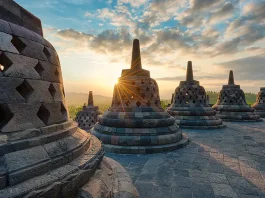 A stunning sunrise view at the ancient Buddhist temple of Borobudur in Java, Indonesia. The photo captures the silhouette of multiple stupas, each adorned with intricately carved stone lattices, against the golden light of the sun peeking through the main stupa. The surrounding landscape of lush mountains under a sky scattered with soft clouds adds to the tranquil and historic ambiance.