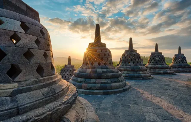 A stunning sunrise view at the ancient Buddhist temple of Borobudur in Java, Indonesia. The photo captures the silhouette of multiple stupas, each adorned with intricately carved stone lattices, against the golden light of the sun peeking through the main stupa. The surrounding landscape of lush mountains under a sky scattered with soft clouds adds to the tranquil and historic ambiance.