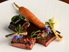 2024 Culinary Travel Trend Predictions Solidify Santa Monica’s Spot at Top of the “Food Chain” A gourmet plated dish featuring medium-rare sliced steak garnished with herbs and edible blue flowers, accompanied by a whole roasted carrot, a charred bone with marrow, and a dollop of puree on a white plate.
