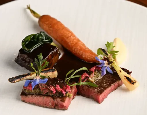 2024 Culinary Travel Trend Predictions Solidify Santa Monica’s Spot at Top of the “Food Chain” A gourmet plated dish featuring medium-rare sliced steak garnished with herbs and edible blue flowers, accompanied by a whole roasted carrot, a charred bone with marrow, and a dollop of puree on a white plate.