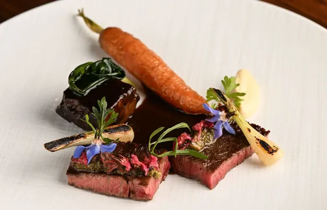 A gourmet plated dish featuring medium-rare sliced steak garnished with herbs and edible blue flowers, accompanied by a whole roasted carrot, a charred bone with marrow, and a dollop of puree on a white plate.