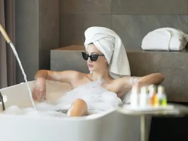 A person with their hair wrapped in a towel and wearing sunglasses is relaxing in a bubble bath inside a bathroom with modern decor. A faucet with running water, toiletries, and folded towels are visible around the bathtub